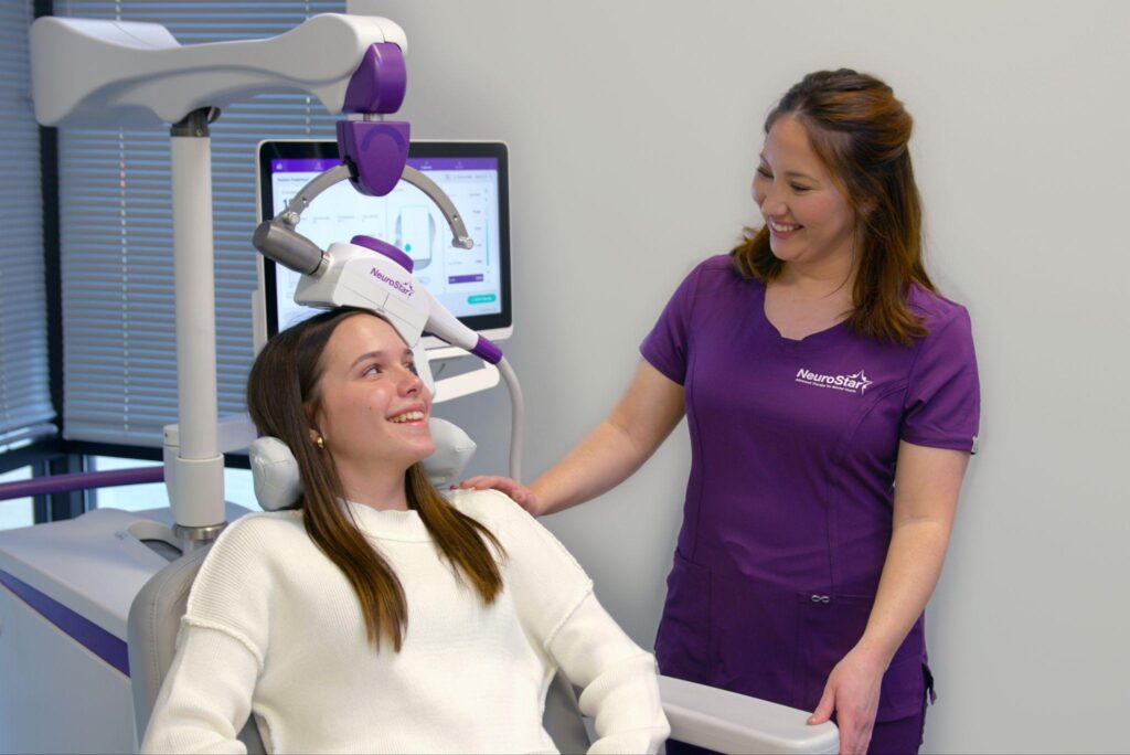Smiling teen girl undergoing NeuroStar TMS treatment while a clinician in purple scrubs stands beside her.